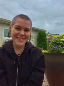 A white woman with short hair smiles at the camera, outside of a house and sitting next to a pot of yellow and purple flowers.