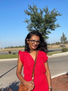 A woman of colour with long dark hair, wearing a red dress, stands outside in front of a tree, and smiles at the camera.